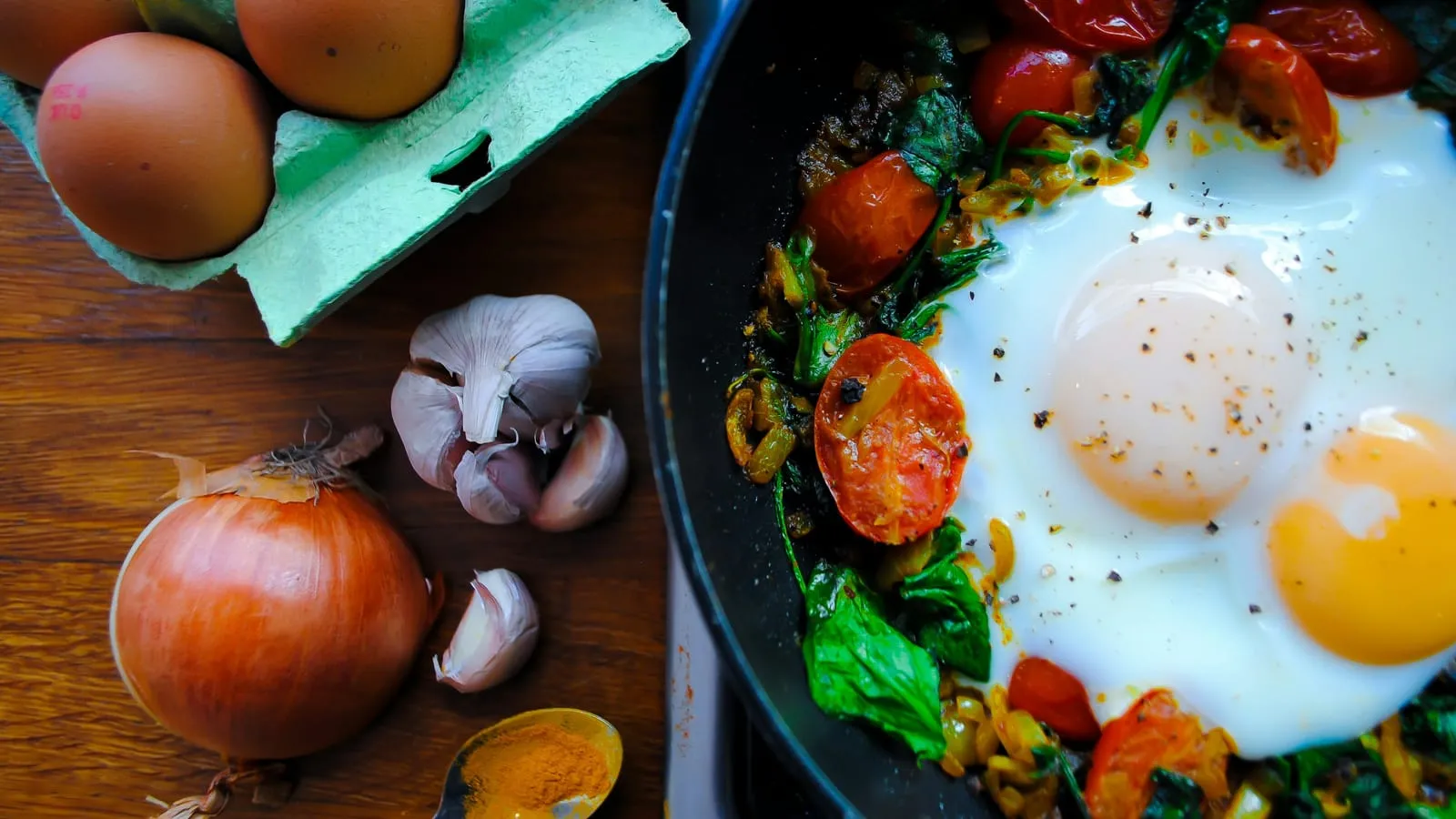 Eggs cooking with spinach and tomatoes in a cast-iron pan, next to an onion, garlic and a carton of eggs