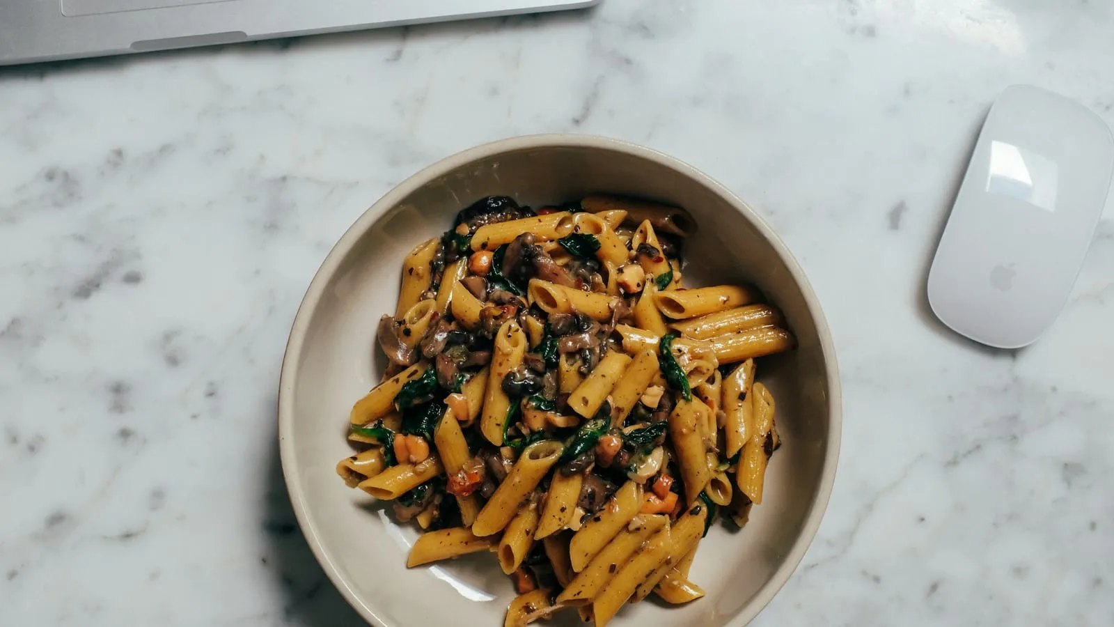 A single bowl of penne with spinach and mushrooms on a marble desk next to a laptop